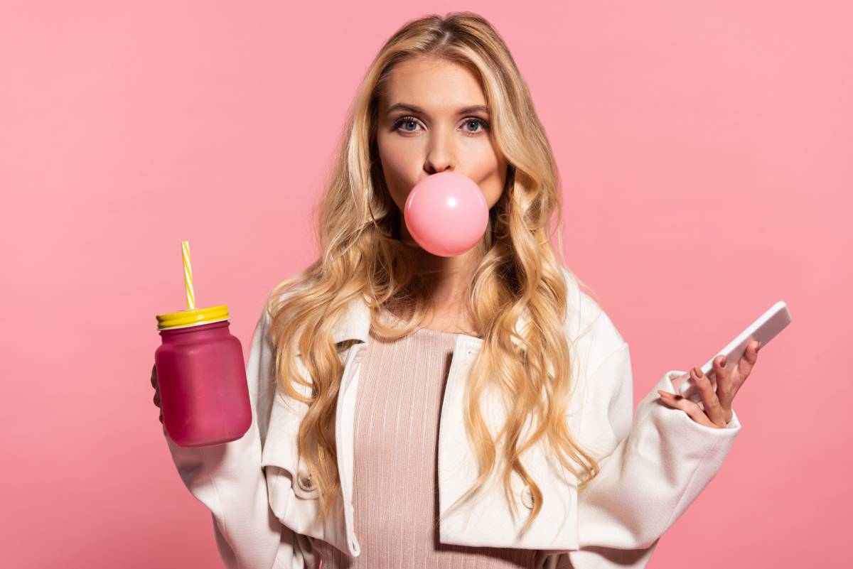 blonde young woman with pink bubble gum in mouth holding smartphone and jar isolated on pink