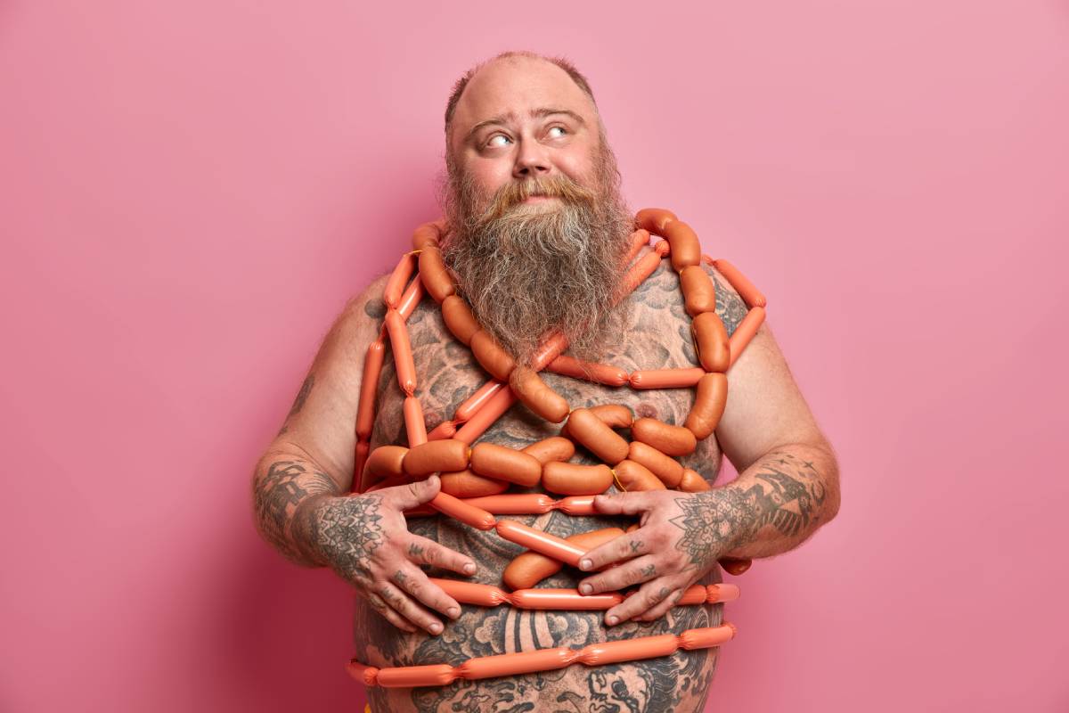 Food addiction and bad nutrition concept. Plump bearded man stands wrapped in sausages has big fat abdomen tattooed body and thick beard leads unhealty lifestyle poses against pink background