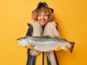 Horizontal shot of positive female angler holds big caught fish wears black raincoat and hat carries fishing net around neck smiles positively isolated over yellow background. Look at my trophy