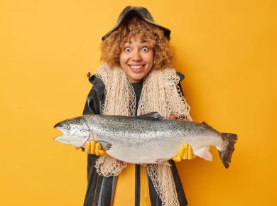 Horizontal shot of positive female angler holds big caught fish wears black raincoat and hat carries fishing net around neck smiles positively isolated over yellow background. Look at my trophy