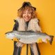 Horizontal shot of positive female angler holds big caught fish wears black raincoat and hat carries fishing net around neck smiles positively isolated over yellow background. Look at my trophy