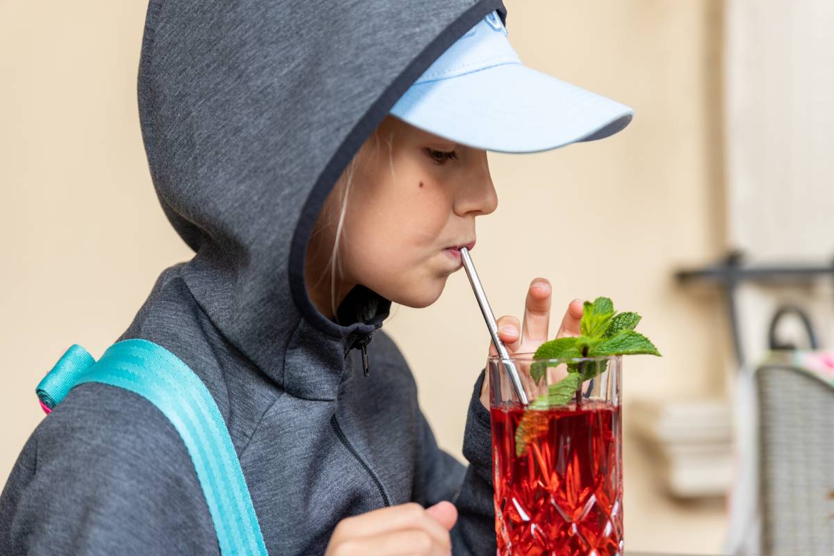 Profile portrait view of cute preteen little kid girl in grey hoodie enjoy drinkong healthy berry lemonade juice compote cocktail walking outdoors. Thirsty child drink beverage outside