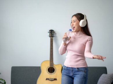 Woman wearing headphones singing with water bottle, standing in living room with a guitar in the background, expressing joy and passion for music.