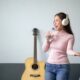 Woman wearing headphones singing with water bottle, standing in living room with a guitar in the background, expressing joy and passion for music.