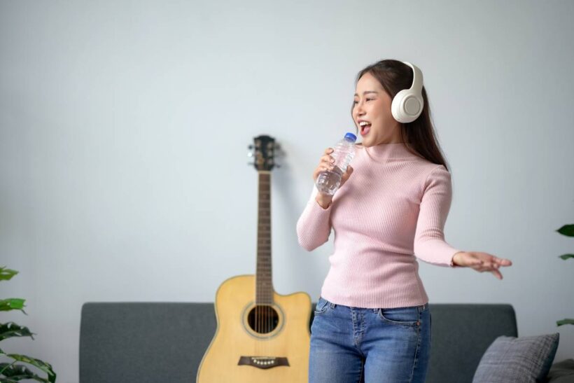 Woman wearing headphones singing with water bottle, standing in living room with a guitar in the background, expressing joy and passion for music.